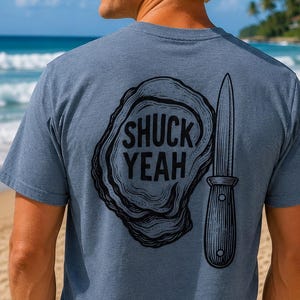 Könnte beinhalten: Ein meliertes blaues Kurzarm-T-Shirt mit einer schwarzen Grafik auf der Rückseite. Das Design zeigt eine Austernschale neben einem Austernmesser, wobei der Text SHUCK YEAH in fetten schwarzen Großbuchstaben in der Schale steht.