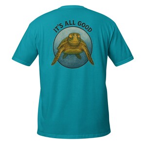 Könnte beinhalten: Türkisfarbenes T-Shirt mit einer detaillierten Illustration einer Meeresschildkröte in einem blauen Kreis. Der Text "IT'S ALL GOOD" steht über der Schildkröte. Das Shirt ist aus weichem Material und hat kurze Ärmel.