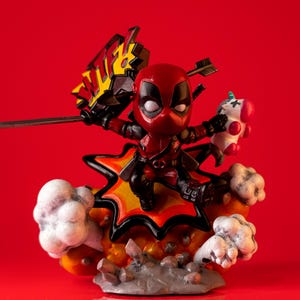 May include: A Deadpool figurine in a dynamic pose, wielding a sword and holding a unicorn plushie. The figure is predominantly red and black, set against a vibrant red background. The base features a stylized explosion with smoke effects.