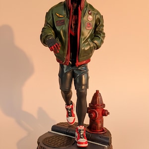 May include: A detailed figurine of Spider-Man Miles Morales in a green jacket, black shorts, and red and white sneakers. The figure is posed on a base with a manhole cover and a red fire hydrant. The base reads "Spider-Man Miles Morales."
