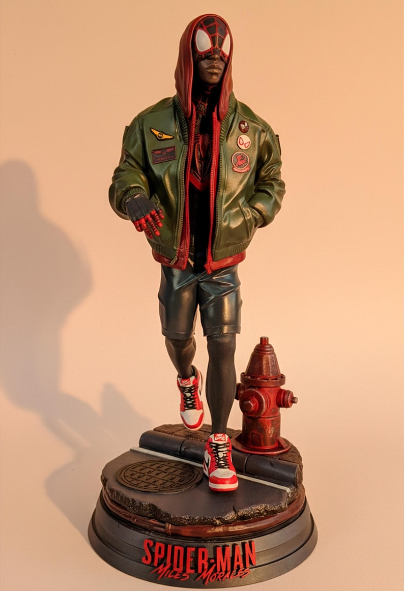 May include: A detailed figurine of Spider-Man Miles Morales in a green jacket, black shorts, and red and white sneakers. The figure is posed on a base with a manhole cover and a red fire hydrant. The base reads "Spider-Man Miles Morales."