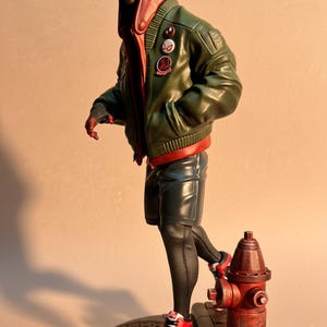 May include: A figurine of a character in a red and black mask, wearing a green bomber jacket, black shorts, and red and white sneakers. The figure is posed near a red fire hydrant.
