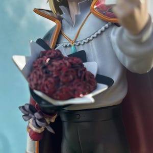 K&ouml;nnte beinhalten: Figur eines Charakters mit blonden Haaren, der ein wei&szlig;es Hemd, eine schwarze Hose und einen roten Umhang tr&auml;gt. Die Figur h&auml;lt einen Strau&szlig; roter Rosen und eine einzelne Rose. Die Figur steht vor einem Hintergrund aus blauem Himmel und gr&uuml;nem Gras.