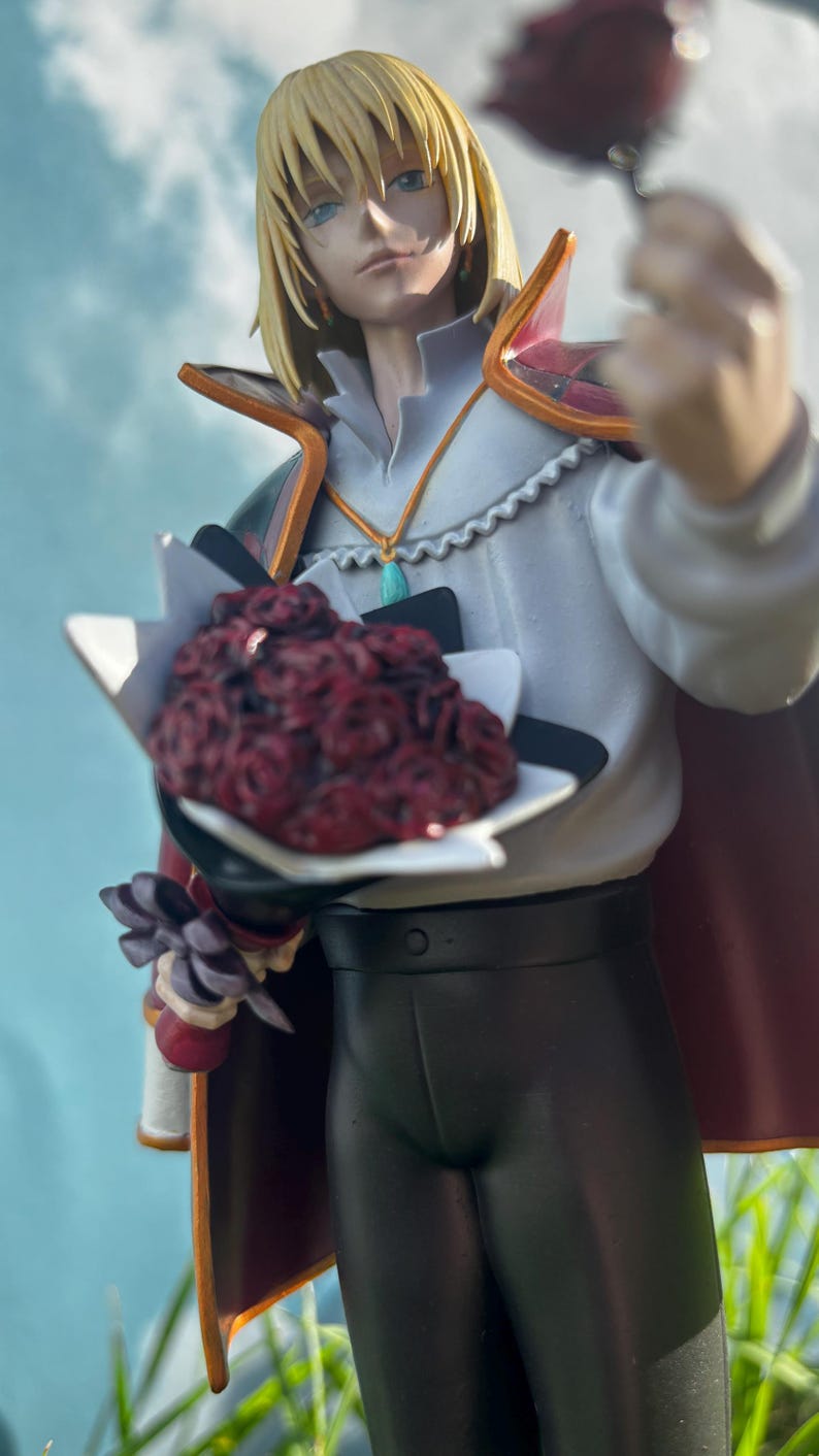 K&ouml;nnte beinhalten: Figur eines Charakters mit blonden Haaren, der ein wei&szlig;es Hemd, eine schwarze Hose und einen roten Umhang tr&auml;gt. Die Figur h&auml;lt einen Strau&szlig; roter Rosen und eine einzelne Rose. Die Figur steht vor einem Hintergrund aus blauem Himmel und gr&uuml;nem Gras.
