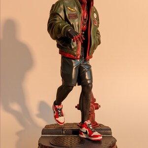 May include: A figurine of Spider-Man Miles Morales in a red and black Spider-Man suit, green bomber jacket, and red and white sneakers. The figure is standing on a base with the words "Spider-Man Miles Morales".