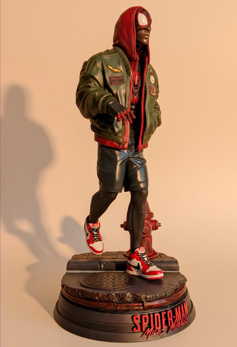 May include: A figurine of Spider-Man Miles Morales in a red and black Spider-Man suit, green bomber jacket, and red and white sneakers. The figure is standing on a base with the words "Spider-Man Miles Morales".