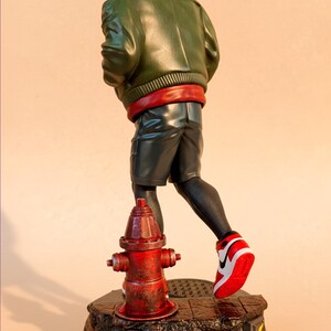 May include: A figurine of a character in a green jacket, red hood, and black shorts, running past a red fire hydrant. The figure is on a black base with a lightning bolt design. The character is wearing red and white sneakers.