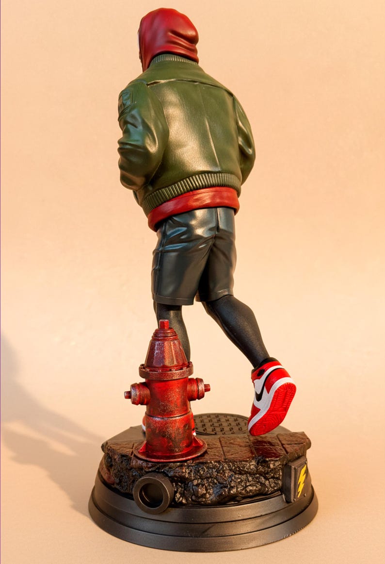 May include: A figurine of a character in a green jacket, red hood, and black shorts, running past a red fire hydrant. The figure is on a black base with a lightning bolt design. The character is wearing red and white sneakers.
