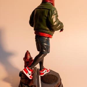 May include: A figurine of a character in a red hoodie and green jacket, black leggings, and blue shorts, wearing red and white sneakers. The figure stands on a base with a fire hydrant and a manhole cover.