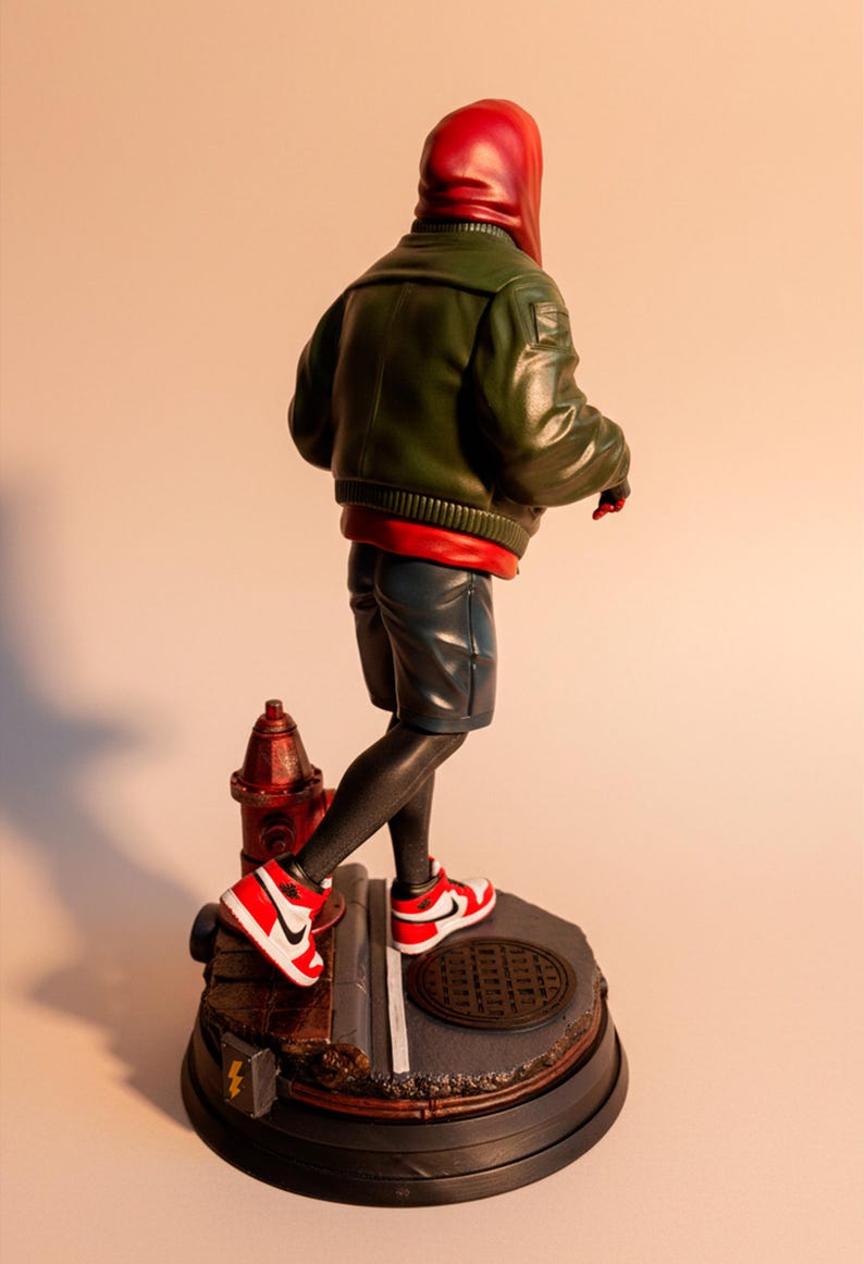May include: A figurine of a character in a red hoodie and green jacket, black leggings, and blue shorts, wearing red and white sneakers. The figure stands on a base with a fire hydrant and a manhole cover.