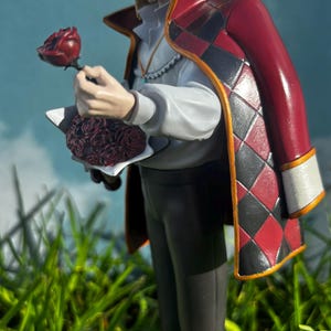 K&ouml;nnte beinhalten: Figur eines Charakters mit blonden Haaren, der ein wei&szlig;es Hemd, eine dunkle Hose und einen roten Mantel mit schwarz-roten Rautenmustern tr&auml;gt. Die Figur h&auml;lt eine rote Rose und einen Rosenstrau&szlig;. Der Hintergrund ist ein blauer Himmel und gr&uuml;nes Gras.