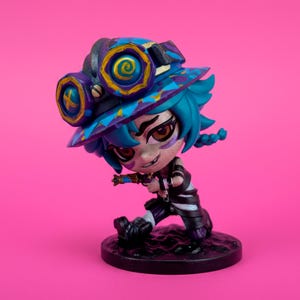 May include: A figurine of a character with blue hair, wearing a hat with goggles, and a black and white striped outfit. The figurine is on a black base, set against a pink background. The hat has a blue, purple, and gold design.