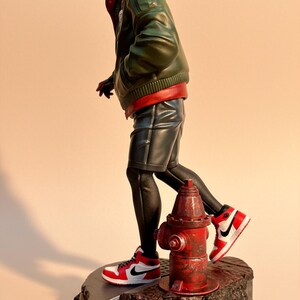 May include: A figurine of a superhero in a red and green outfit, black shorts, and red and white sneakers. The figure is standing on a base with a red fire hydrant. The superhero is wearing a red hood and a green jacket.