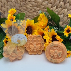 May include: An arrangement of honey-themed soaps and a small bag of soaps, decorated with sunflowers. The soaps are orange and shaped like a honeycomb and a circle with a bee design. The bag has a tag that says "Thank you for coming!"