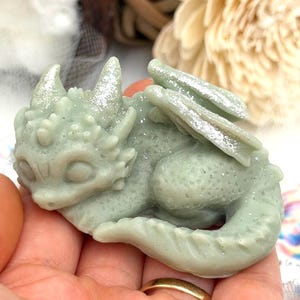 May include: A light green dragon figurine with glitter accents on its horns and wings. The dragon is in a curled-up pose, with detailed scales and a friendly expression. The figurine is held in a hand, showcasing its size and form.