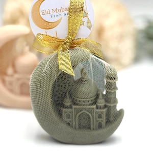 May include: A decorative soap shaped like a crescent moon with a mosque design, wrapped in a green mesh and tied with a gold ribbon. A round tag with the text "Eid Mubarak" is attached.