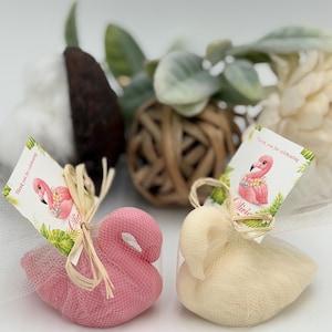 May include: Two swan-shaped bath sponges, one pink and one cream, each wrapped in white netting and tied with a raffia bow. Attached to each is a rectangular tag featuring a pink flamingo illustration and the text Thank you for celebrating.
