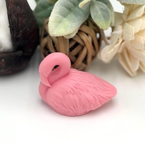 May include: A small, pink flamingo figurine with detailed feather texture. The flamingo is resting on a white surface, with decorative elements including a woven ball, faux greenery, and a cream-colored flower in the background.