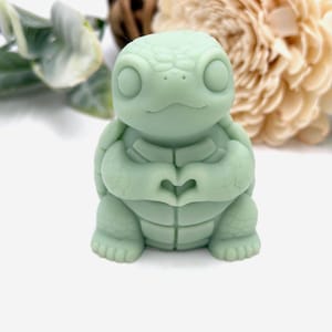 May include: A light sage green turtle figurine with a smooth matte finish. The turtle is depicted in a seated position with its front flippers positioned together in front of its chest to form a heart shape. It has large, round, blank eyes.