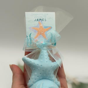 May include: A light blue starfish-shaped item is encased in a white mesh bag, tied with a light blue ribbon. A small card with the name "JAMES" and a starfish graphic is attached. The overall theme suggests a nautical or beach-themed party favor.