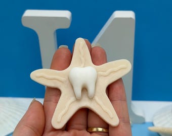 May include: A beige starfish-shaped object with a white tooth in the center. The starfish has textured lines and is held against a blue background with white letters.