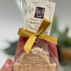 May include: A graduation-themed gift set in a clear cellophane bag, tied with a gold ribbon. The set includes a light-colored soap with "Congrats GRAD" embossed on it and a small card with a photo and text.