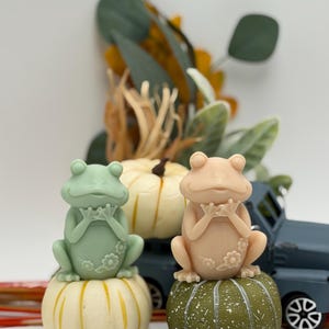 May include: Two frog figurines, one mint green and one peach, sit atop decorative pumpkins. The frogs have their hands clasped and are adorned with floral details. A miniature blue truck and fall foliage are in the background.