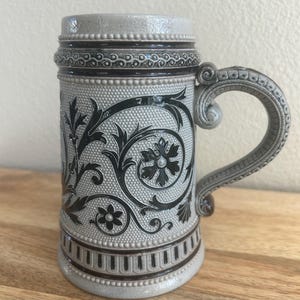 May include: A grey stoneware beer stein featuring a dark brown relief pattern of scrolling acanthus leaves and floral motifs. The cylindrical vessel has a textured stippled background, a decorative beaded rim, and a curved handle with ornate detailing.