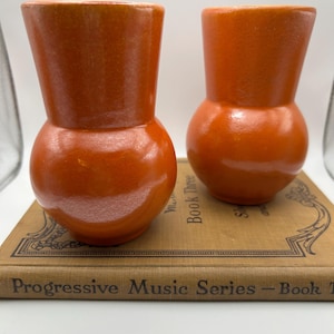 May include: Two orange ceramic vases with a bulbous base and flared top. The vases are placed on a vintage book with the text "Progressive Music Series - Book Three". The vases have a glossy finish and a warm, inviting color.