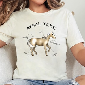 May include: Cream-colored t-shirt featuring a graphic of an Akhal-Teke horse with the text "AKHAL-TEKE" above it. The horse is tan with a white mane and tail. The shirt also has the words "Handy", "Agile", "Intelligent", "Loyal", and "Strong" around the horse.