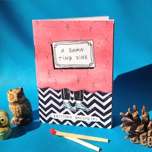 May include: A pink and red illustrated zine with the title "A Damn Tiny Zine" in a white frame. The zine is sitting on a black and white chevron pattern. The text "KRYSTYNA BACZYNSKI" is written in white at the bottom of the zine.