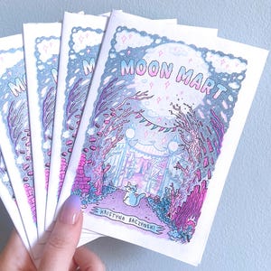 以下が含まれることがあります： 小さな白いジンが複数枚重なっている。表紙にはピンク色の「MOON MART」の文字、猫が登場する空想的なシーン、アーティスト名が描かれている。背景は水色で、ジンは手で持たれている。