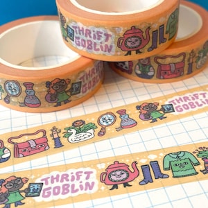 May include: Three rolls of orange washi tape with the text "Thrift Goblin" and various illustrations. The tape features images of a teapot, boots, a doll, and other whimsical designs. The tape is on a white and blue grid background.