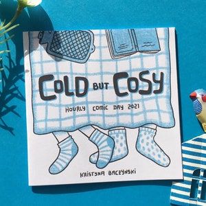 May include: A white comic book with a blue and white checkered pattern on the cover. The title is "COLD BUT COSY" with the words "HOURLY COMIC DAY 2021" below. The author's name is "KRYSTYNA BACZYNSKI" at the bottom.