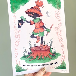 May include: A rectangular art print featuring a dark-skinned character with a straw hat, wearing a tunic and pointed shoes, standing on a tree stump. The character holds a lantern and a flute. The text reads, NOT ALL THOSE WHO WANDER ARE LOST.