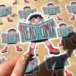 May include: A die-cut sticker featuring a cartoon character with short black hair, wearing a black outfit and red boots, holding a large light blue book open. The book cover displays the red text READ MORE COMIX in a bold, stylized font.