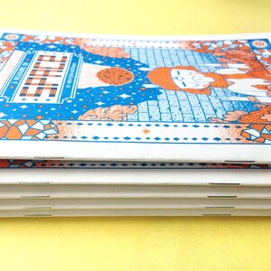 May include: A stack of four small, white, bound books with orange and blue covers. The cover features a colorful illustration of a person with a red beard and a blue background with stars. The title of the book is "A Leisure of Stitch" by Kristin Nicholas.