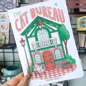 May include: A rectangular art print titled THE CAT BUREAU featuring a green and white building with a balcony, a red door, and a cat sitting in a chair reading a newspaper. The illustration includes a street lamp, a tree, and the text ENTER HERE.
