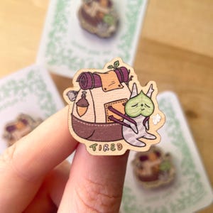 May include: A wooden pin featuring a cartoon creature with a green leaf mask, carrying a tan and purple backpack with an acorn charm. The creature is sitting down, and the word TIRED is printed in colorful letters at the bottom of the pin.
