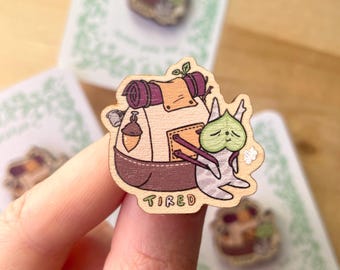May include: A wooden pin featuring a cartoon creature with a green leaf mask, carrying a tan and purple backpack with an acorn charm. The creature is sitting down, and the word TIRED is printed in colorful letters at the bottom of the pin.