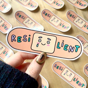 May include: A peach-colored sticker in the shape of a bandage with the word "RESILIENT" in teal and purple. The bandage has a cartoon face with a tear. Other stickers with the same design are in the background.