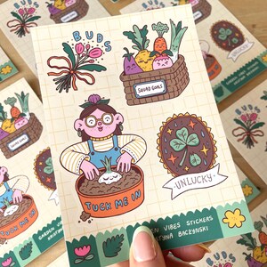 May include: A sheet of colorful stickers with a garden theme. Designs include a basket of vegetables, a gardener, a four-leaf clover, and the text "Tuck Me In" and "Squad Goals". The stickers are on a grid-patterned background.