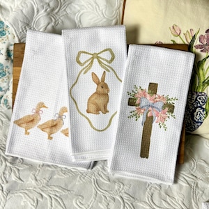 May include: Three white waffle-weave kitchen towels with Easter-themed designs. One towel features three ducks with bows, another has a bunny in a gold frame, and the third displays a cross adorned with flowers and a bow.