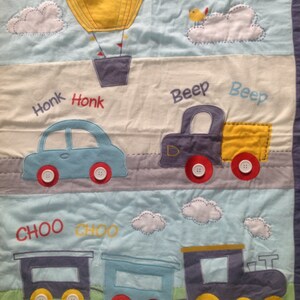 May include: A blue and yellow baby quilt with a hot air balloon, two cars, a train, and clouds. The quilt has the words "Honk Honk", "Beep Beep", and "Choo Choo" embroidered on it.