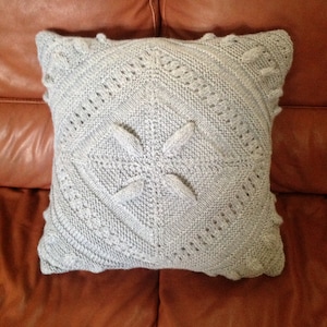May include: A gray knitted pillow with a geometric pattern. The pillow is on a brown leather couch.