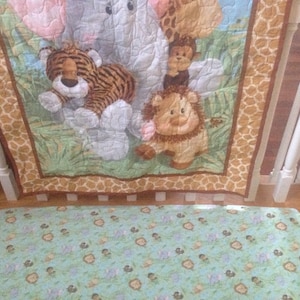 May include: A baby crib bumper with a jungle animal theme. The bumper features a brown and white elephant, a tiger, a lion, and a monkey. The fabric is a light brown with a giraffe print.