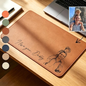 Op de afbeelding: Een rechthoekige, bruin lederen bureaumat met de tekst "I love you, Daddy" en een schets van een vader en kind. Een kleine foto van de vader en het kind staat in de rechterbovenhoek. Een notitieblok, pen en koffiekopje zijn ook zichtbaar.