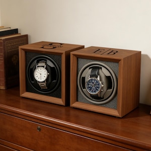 May include: Two wooden watch winders with watches inside sit on a wooden chest of drawers. The winders are square with a dark interior and a watch holder. One watch has a silver band and white face. The other has a black band and blue face.