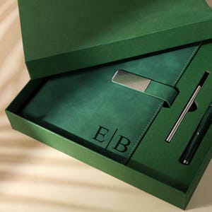 May include: A green gift set in a green box. The set includes a green leather notebook with a silver metal clasp, a pen, and a silver metal pen. The notebook is embossed with the initials "E/B".