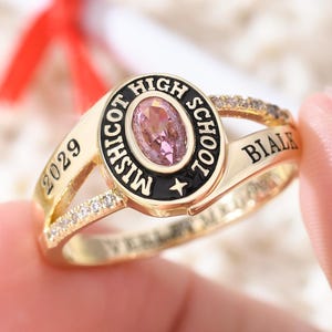 May include: A gold-colored class ring with a pink oval-shaped stone at the center. The ring features the words "Mishicot High School" and "2029". The ring is held by a person's fingers.